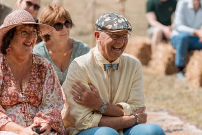 Lachende gasten met zomerhoeden en een vlinderdas zitten op strobalen tijdens de bruiloft van Bert en Verena
